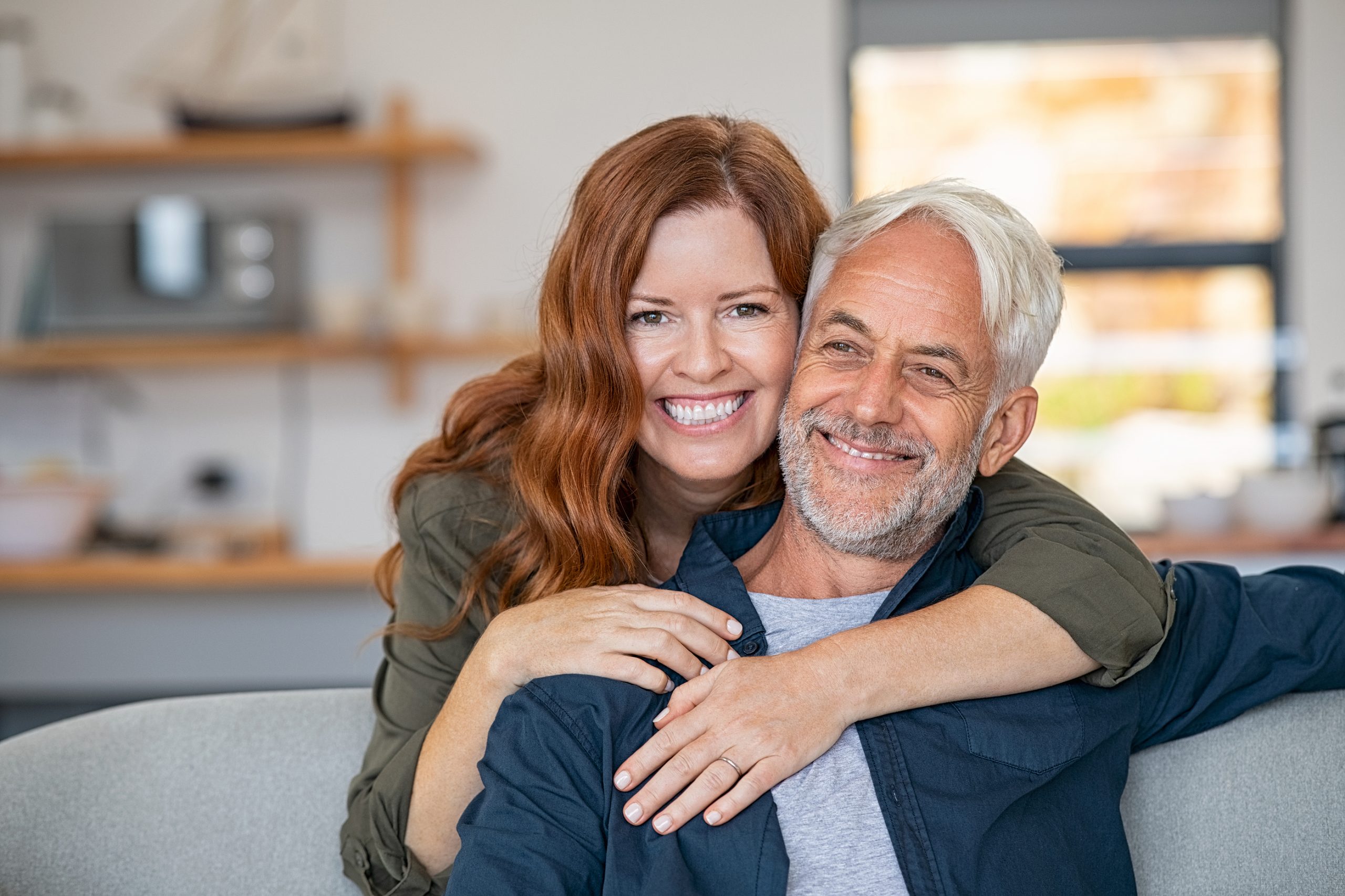 Smiling woman hugging her senior husband on couch from behind in