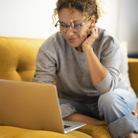 Happy Young Woman Working On Laptop Sitting On Sofa In Living Ro