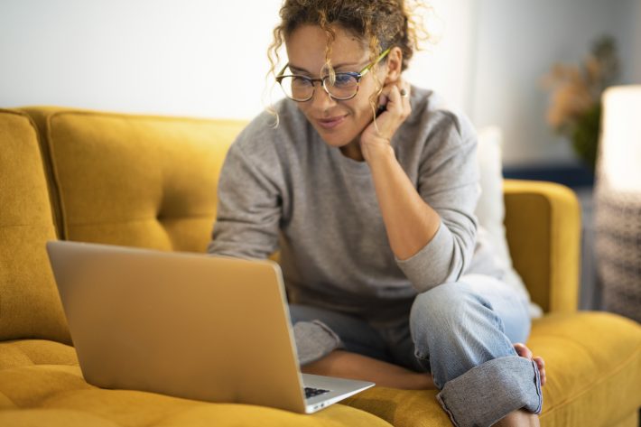 Happy Young Woman Working On Laptop Sitting On Sofa In Living Ro
