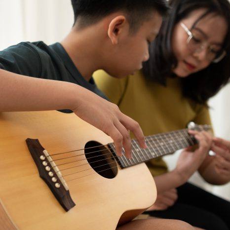 Asian Boy Playing Guitar With Mom In The Living Room For Teachin