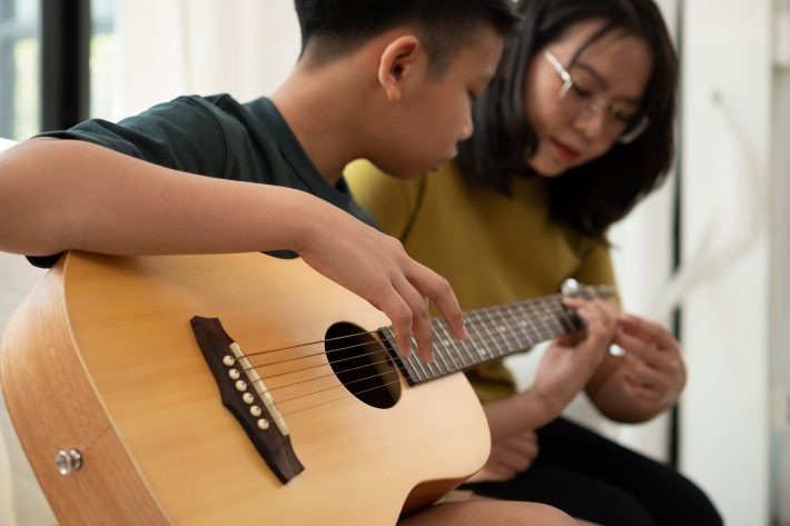 Asian Boy Playing Guitar With Mom In The Living Room For Teachin
