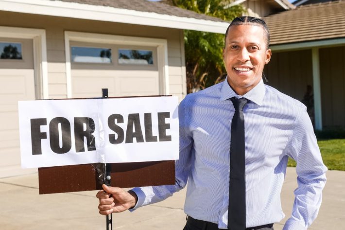 Smiling real estate agent with a for sale sign in front of a house